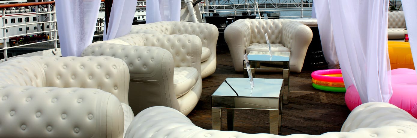A luxurious outdoor lounge features white, plush, tufted sofas and a mirrored coffee table on a wooden deck. Sheer white curtains frame the seating area, while inflatable event furniture adds a touch of whimsy. In the background, a pool float and part of a ship complete the serene scene.
