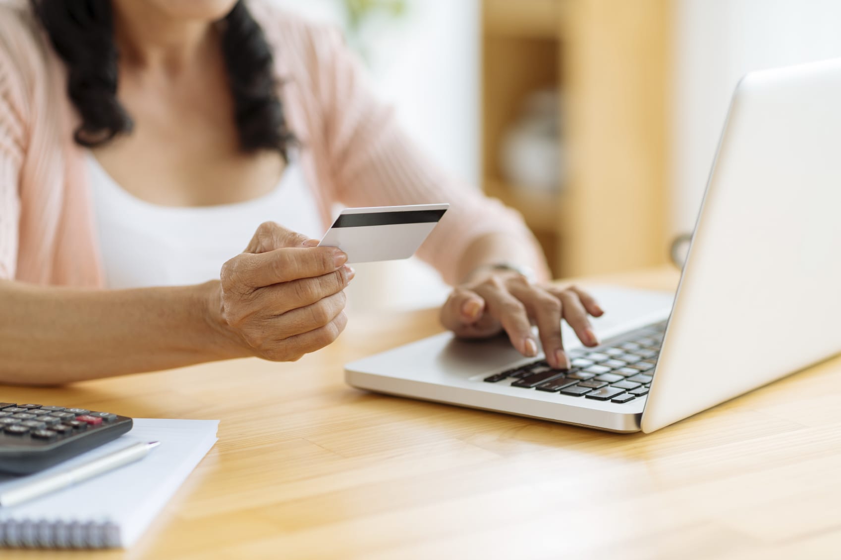A person at a wooden table, partially shaded by a custom canopy, holds a credit card in one hand while typing on a laptop with the other. Nearby are a notebook, a pen, and a calculator.