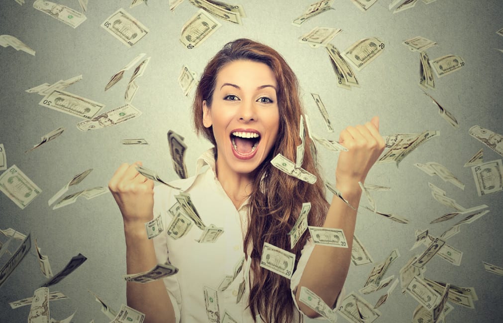 A woman with long hair is smiling and cheering with her fists raised, surrounded by floating dollar bills against a plain background. She appears joyful and excited, as if she's just hit the jackpot at one of those thrilling expo shows.