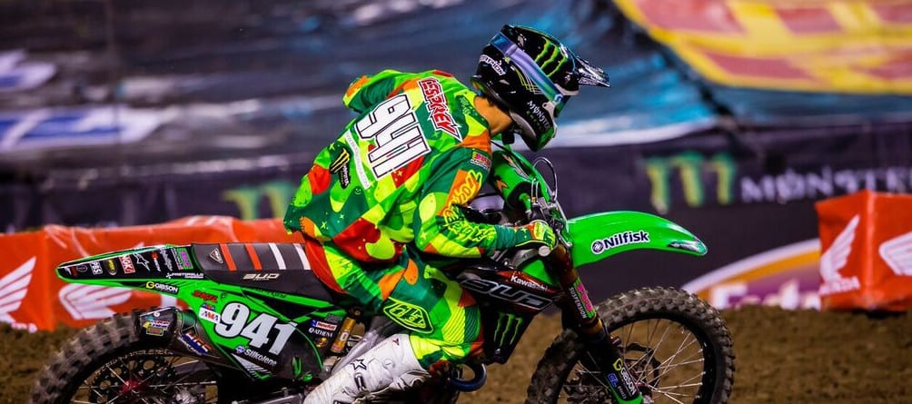 A motocross rider in a vibrant green, red, and yellow outfit navigates a dirt track on a green motorcycle, number 941 prominently displayed. Event custom tents line the track, their banners and lights creating a dynamic backdrop for the thrilling ride.