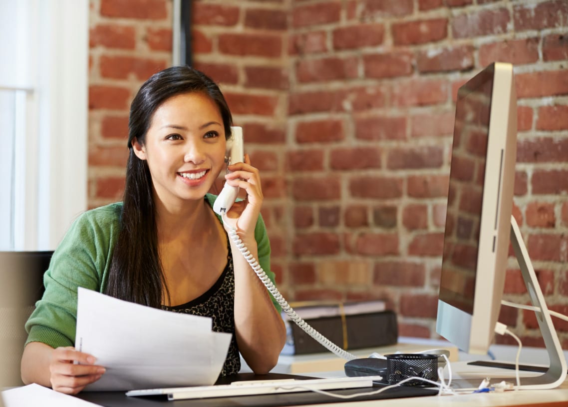 A woman sits at a desk, smiling as she talks on a corded telephone with papers in her other hand. A computer monitor is in front of her, while the backdrop reveals a brick wall adorned with colorful illustrations of pop-up tents.