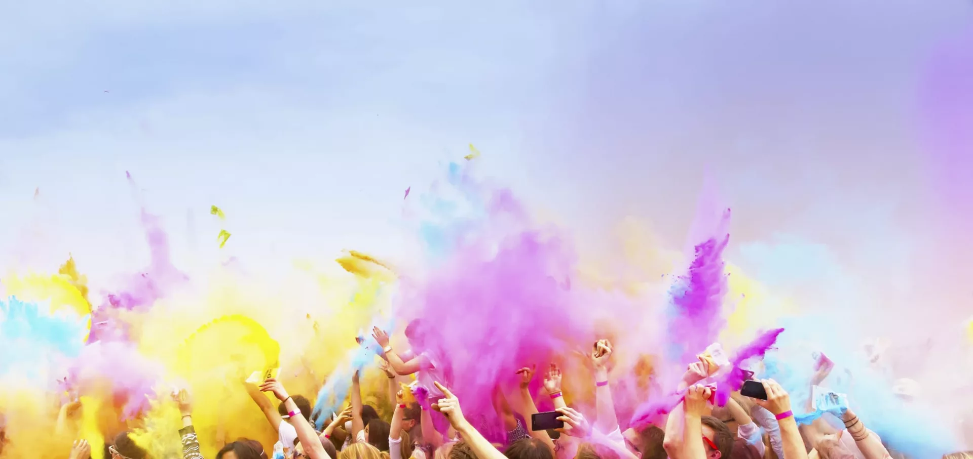 A crowd of people celebrates by throwing vibrant colored powders into the air. Beneath a clear sky, a custom tent provides shade while the mix of yellow, pink, and purple colors creates a festive and joyful atmosphere.