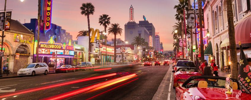 A vibrant city street at dusk is alive with illuminated signs, palm trees, and busy traffic. Neon lights from storefronts and cars illuminate custom printed tents lining the sidewalks, creating a lively scene under a colorful sky.