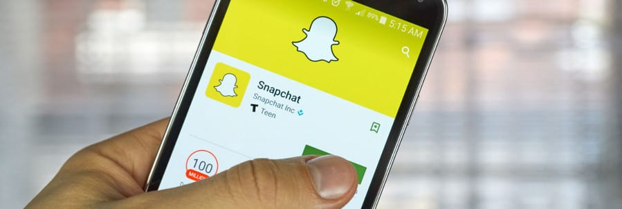 A hand holding a smartphone displays the Snapchat app page on the Google Play Store, showcasing the iconic logo and app details with an install option. It's as inviting as spotting a brightly colored custom tent at a bustling festival.