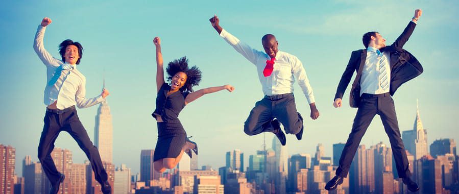 Against a vibrant city skyline, four businesspeople in formal attire leap joyfully, their enthusiasm reminiscent of a successful trade show booth. The clear sky and shining sun amplify the energy and positivity, with skyscrapers like the Empire State Building standing tall in the background.