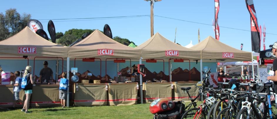 A line of commercial pop-up tents adorned with Clif logos showcases various products at an outdoor event. People browse the stands, while bicycles rest on a grassy field under a sunny sky. Flags and trees create a picturesque backdrop in the distance.