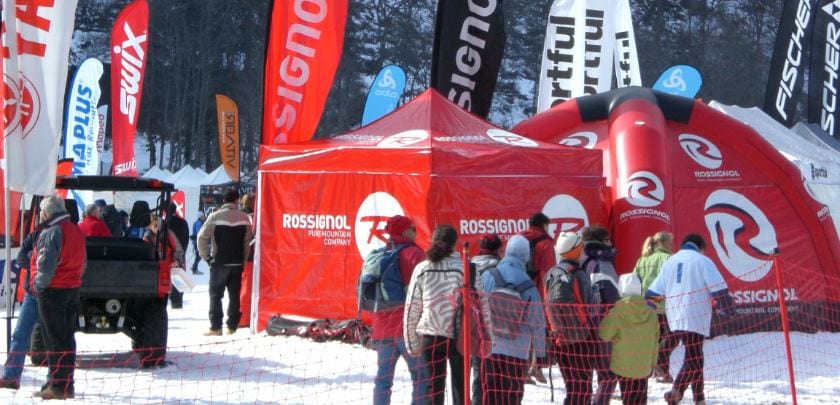 People dressed in winter clothing gather around a Rossignol tent surrounded by branded flags and promotional items on a snowy landscape. A black vehicle is parked nearby, and trees are visible in the background.