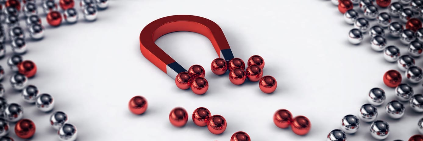 A red horseshoe magnet attracts numerous red metallic spheres, with silver ones scattered around on a white surface, showcasing an innovative way to display promotional products.