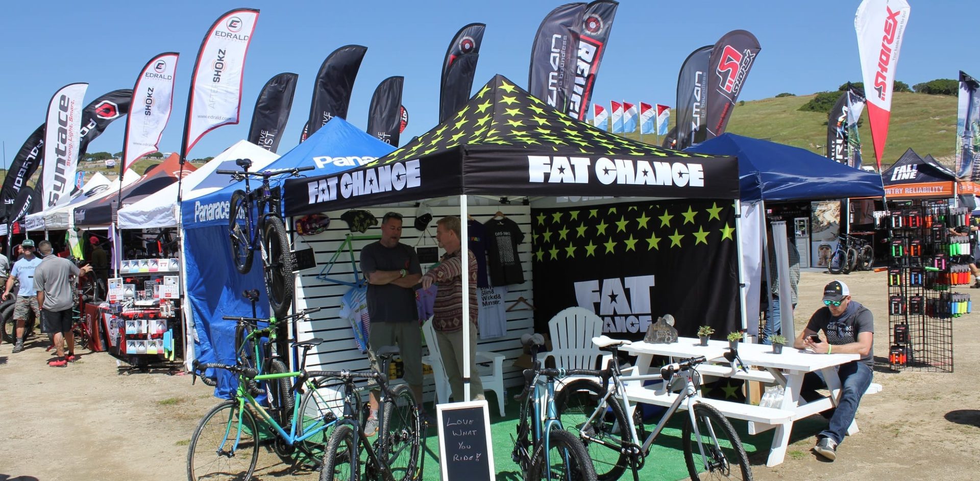 Outdoor bike expo with a Fat Chance booth displaying bicycles under a star-patterned canopy. Several people are standing nearby. Various brand tents and flags are in the background. A person sits at a table on the right.