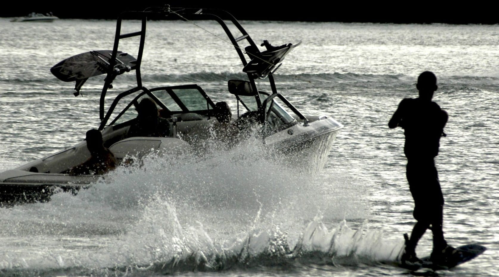 A person wakeboarding is silhouetted in front of a speeding motorboat, which creates a splash and wake on the shimmering water. Tents dot the shoreline, nestled among trees in the background as the sun reflects off the surface.