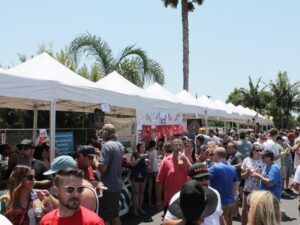 A large crowd of people enjoying a sunny day at an outdoor festival. Several white tents are set up with vendors and activities. Palm trees can be seen in the background, and people are dressed in casual summer clothing.