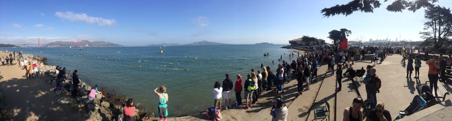 A crowd gathers along a coastal promenade on a sunny day. With Instent excitement, swimmers approach the shore from the bay, and the Golden Gate Bridge is visible in the distance. The sky is clear, creating a vibrant and lively atmosphere.