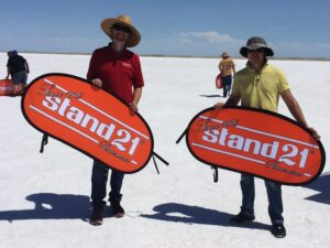 Two people standing on a vast, flat salt surface, holding large orange shields with the text Stand 21. They are surrounded by a few others wearing hats, under a clear blue sky.