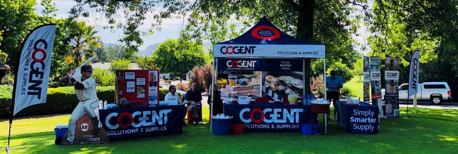 At the outdoor marketing event for Cogent Solutions & Supplies, a custom tent shelters a person surrounded by promotional displays and materials. A life-sized cardboard cutout of a baseball player adds flair, while trees and grassy areas provide a picturesque backdrop. Banners complete the vibrant scene.