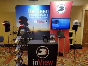 A trade show booth for inView displays wireless brake and turn signal lights for helmets. The booth features a backlit logo, helmets with signal lights, promotional materials, and an information screen on a red backdrop.