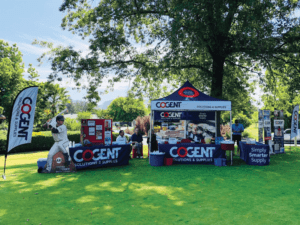 Outdoor promotional setup featuring Cogent Solutions & Supplies. A tent displays products, banners, and signs. A life-size baseball player cutout stands next to the booth. Trees and grass in the background create a park-like setting.