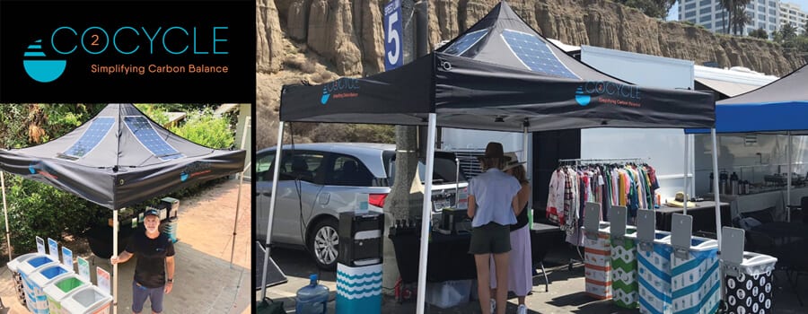 Outdoor market stall for C2Cycle with a black canopy displaying their logo and slogan Simplifying Carbon Balance. Bins for sorting waste and racks of colorful clothing are visible under the tent. Cliffside and parked vehicles in the background.