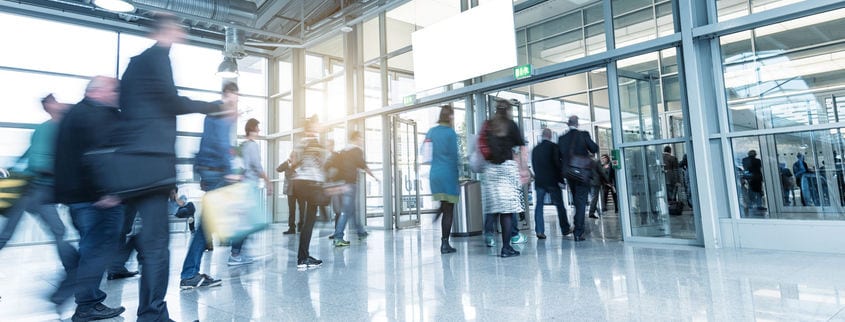 A busy airport terminal with a blurred crowd of travelers moving in various directions. People carry luggage and bags, passing through glass doors under bright overhead lights. A large sign hangs in the center above the entrance.