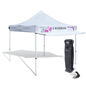 A 10’x10′ Pop Up Canopy Tent with a 10′ awning featuring a 3 Rosebuds Jewelry banner and floral design. The tent is fully set up on a flat surface, with its carrying case and stakes positioned near one of the legs.