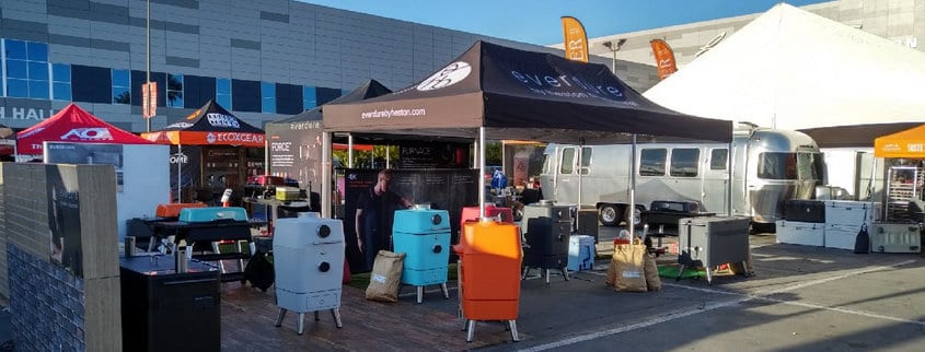At a bustling trade show, vibrant grills and smokers are featured under branded tents with fire certificates for canopies. A gleaming silver Airstream trailer stands in the backdrop against a large exhibition building, while scattered visitors enjoy the sunny weather.