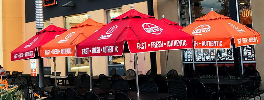 The outdoor dining area features custom commercial umbrellas in vibrant red, each showcasing the words Fast, Fresh, and Authentic. Beneath these striking canopies are black metal tables and chairs, set invitingly outside the building.