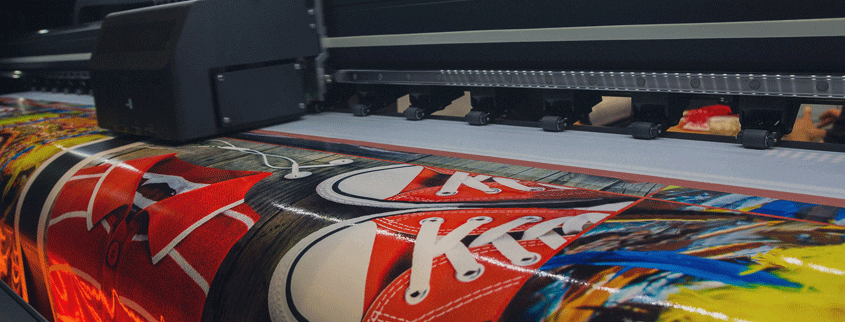 A large dye-sublimation printer producing vibrant, colorful images on a roll of paper, featuring illustrations of red sneakers and abstract designs. The printing process shows high detail and clarity.