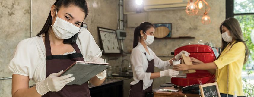 Three women, exemplifying how consumer behavior has changed due to Covid-19, wear face masks and gloves in a coffee shop. One holds a tablet, another prepares a drink, and the third serves a customer. Dressed in white shirts and brown aprons, they stand against modern decor.