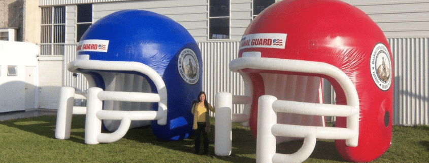 A person stands between two giant inflatable football helmets, one blue and one red, positioned on the grass outside a building.