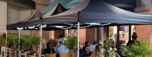 Outdoor dining area with several people seated under black canopy tents adorned with string lights. The setting features brick walls and greenery, creating a cozy atmosphere.