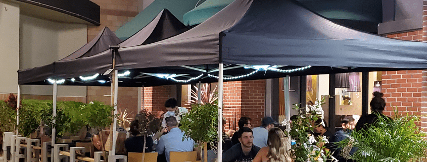 Outdoor dining area with several people seated under black canopy tents adorned with string lights. The setting features brick walls and greenery, creating a cozy atmosphere.