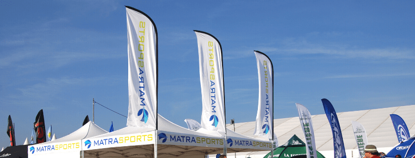 The outdoor event unfolds under a clear blue sky, with flags prominently displaying MATRA SPORTS flanking a branded tent. Additional small tents and vibrant flags showcasing various logos dot the background, creating a lively and colorful atmosphere.
