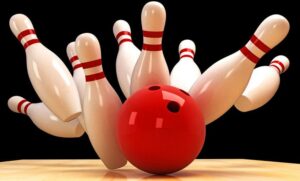 A red bowling ball striking multiple white bowling pins with red stripes, causing them to scatter against a black background. The scene suggests a powerful throw, capturing the moment just before the pins fall completely.