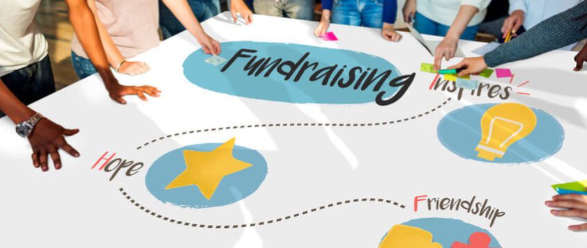 A group of diverse people stands around a large paper on a table with the word Fundraising in the center. Surrounding words include Inspires, Hope, and Friendship, with drawings of a lightbulb and a star.