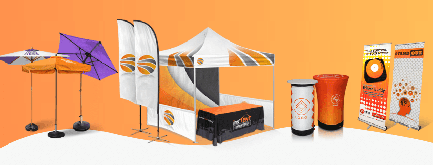 A range of branded display items on an orange background, including promotional tents, umbrellas, banners, a table cover, a counter, and a roll-up stand. The items feature matching orange, black, and white designs with abstract logos.
