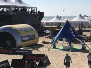 A beach event featuring several tents and promotional booths, with people walking around on the sand. A large inflatable structure and branded banners are visible. The ocean and kite surfers are in the background.