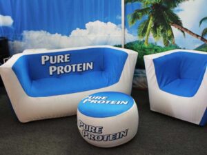 A furniture set with a couch, chair, and ottoman is covered in blue and white fabric, branded with the words Pure Protein. A backdrop features a tropical scene with a palm tree and blue sky.