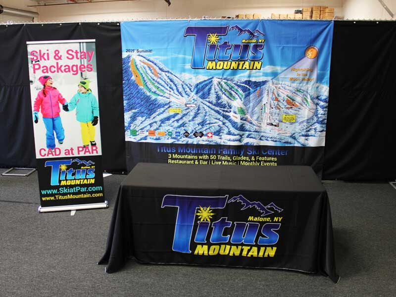 A promotional display for Titus Mountain in Malone, NY, featuring a detailed map of the ski resort, a banner on the table reading Titus Mountain, and a vertical sign advertising ski and stay packages.