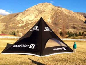 A person stands next to a large, black, triangular tent with Salomon logos, set against a backdrop of a rocky hill under a blue sky. The tent is pitched on grassy terrain, with a white fence visible in the background.