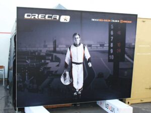 A large poster featuring a race car driver in racing gear holding a helmet, set against a racetrack background. The top left displays the ORECA logo, and digital numbers are shown to the right.