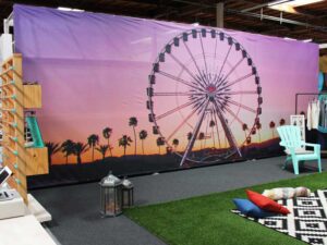 A room features a large mural of a sunset scene with a Ferris wheel and palm trees. Theres a turquoise chair, colorful cushions, and a small rug on the floor. A lantern and displays with clothes add to the decor.