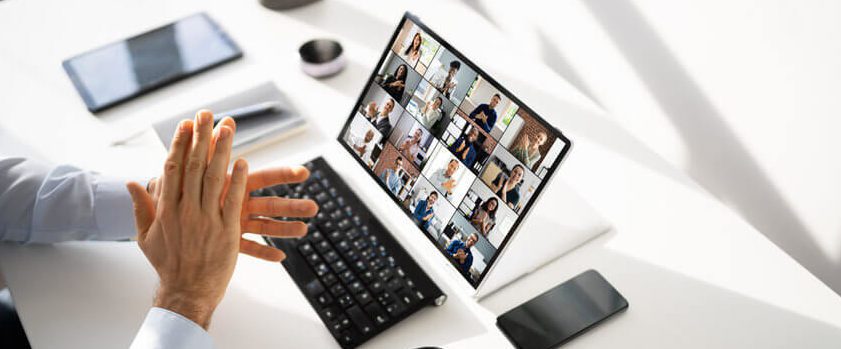 A person sits at a desk with a laptop displaying a grid of people in a video conference. Their hands are clasped in front, and a keyboard, smartphone, and coffee cup are nearby. Bright lighting streams in from the side.