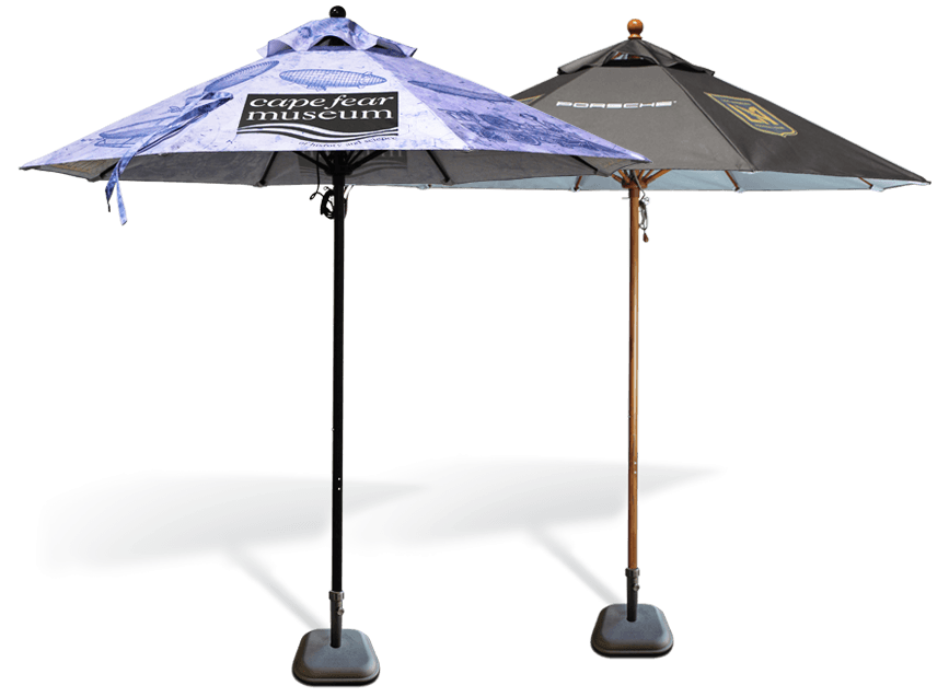 Two large outdoor umbrellas with weighted bases. The left umbrella is blue with a Cape Fear Museum logo. The right umbrella is black with a Porsche logo. Both umbrellas are open and casting shadows.