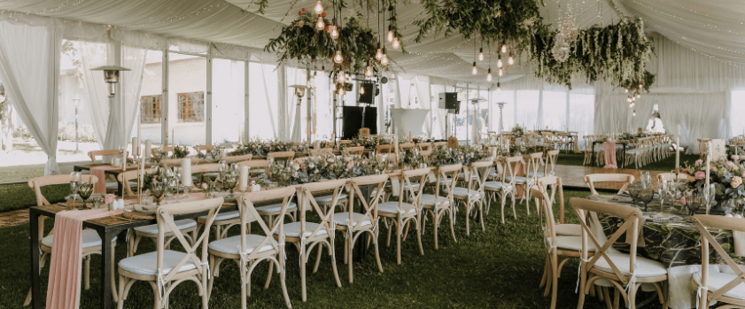 A spacious, elegantly decorated wedding reception under a white canopy tent. Tables are adorned with greenery, flowers, and candles. Wooden chairs with white cushions are neatly arranged, while string lights and hanging foliage add a warm, festive ambiance.