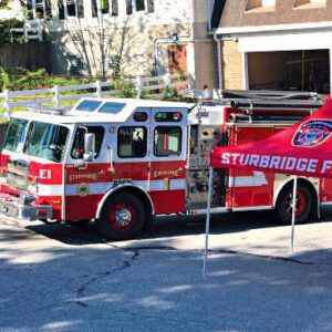 A fire truck labeled Sturbridge Engine 1 parked in front of a building. Beside it, a red tent with Sturbridge Fire Department logo provides shade. Sunny day with shadows on the pavement.