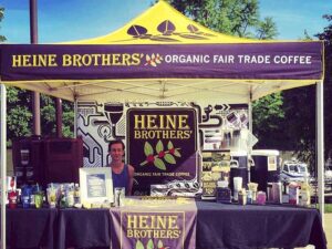 A Heine Brothers Organic Fair Trade Coffee booth is set up at the event, featuring food concession tents. A person stands behind a table with coffee products under a branded tent, surrounded by trees and a blue sky backdrop.