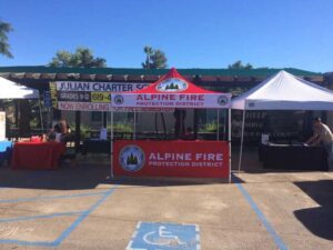 Outdoor event with tents for Alpine Fire Protection District and Julian Charter School. The Alpine Fire tent is red and centrally placed. People are visible under the tents. A sunny day with trees in the background.