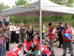A group of emergency responders in red vests attend to a situation under a tent near a body of water. Bystanders, including children and adults, observe from a distance. Photographers capture the scene. Trees and a railing are in the background.
