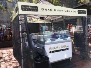 At an outdoor event, a Gnam Gnam Gelato stand nestles under the cozy cover of food concession tents. The display case boasts various gelato containers, with brand signs in plain view. In the background, people enjoy their treats, creating a lively atmosphere.