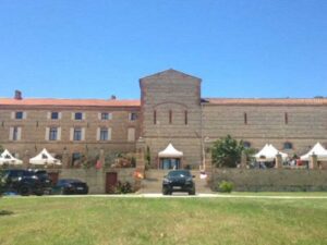A large stone building with multiple windows stands under a clear blue sky. Two black cars are parked in front, and several white tents are set up on the terrace. A manicured lawn fills the foreground.