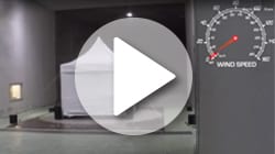 An empty room features a high peak pop-up tent in the center, surrounded by four black weights. A circular gauge on the right shows wind speed at a high level. A play button overlay suggests this is a video thumbnail.